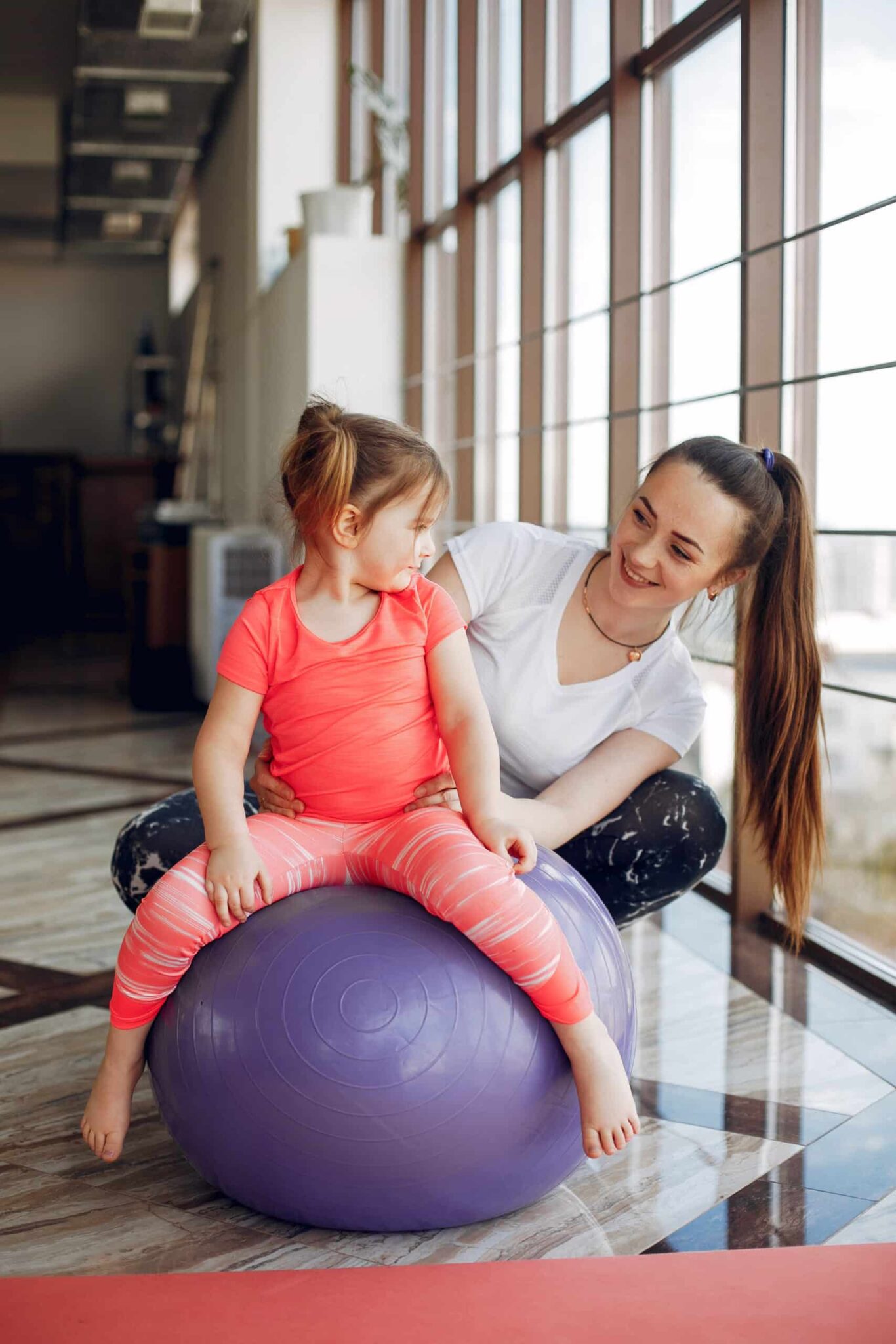 Eine junge Frau mit schulterlangem Haar sitzt hockend in einer schwarzen Fitnesshose und weißem T-Shirt hinter einem jungen Mädchen, das auf einem lilafarbenen Ball mit ca. 60 cm Durchmesser sitzt. Beide schauen sich glücklich an. Das Mädchen trägt ein rotes Sporttrikot und ein pinkes T-Shirt. Der Raum ist hell mit großen Fenstern und wirkt wie ein Therapieraum für Physiotherapeuten.