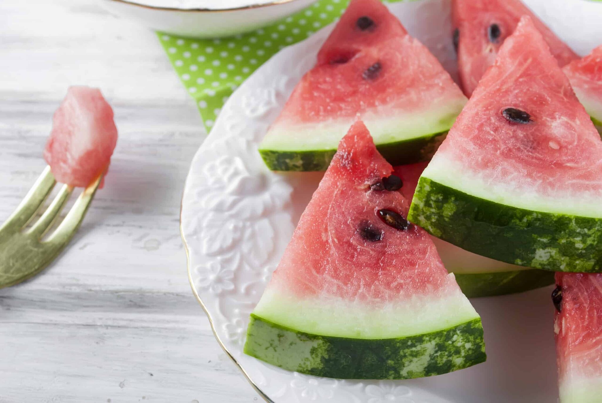 Eine Schüssel mit Wassermelonenkernen auf einem Holztisch. Die Kerne sind schwarz und haben eine ovale Form. Im Hintergrund sieht man eine Wassermelone mit grüner Schale und rotem Fruchtfleisch.