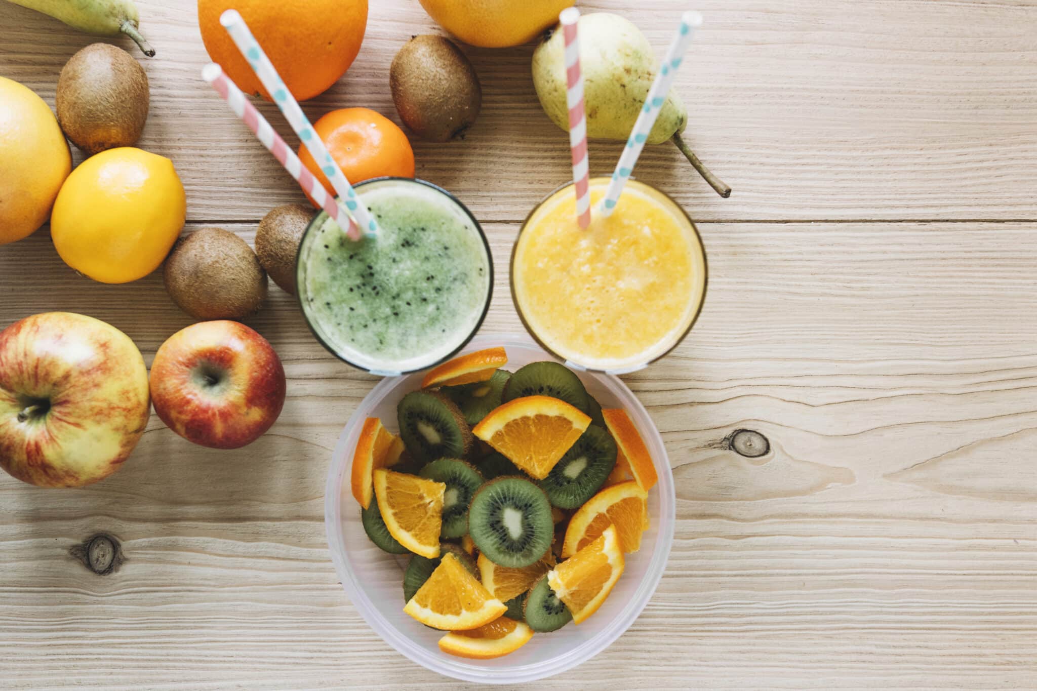 Zwei bunte Smoothies in Glasflaschen und eine Schale mit frischem Obst stehen auf einem Holztisch. Im Hintergrund ist eine grüne Pflanze zu sehen.